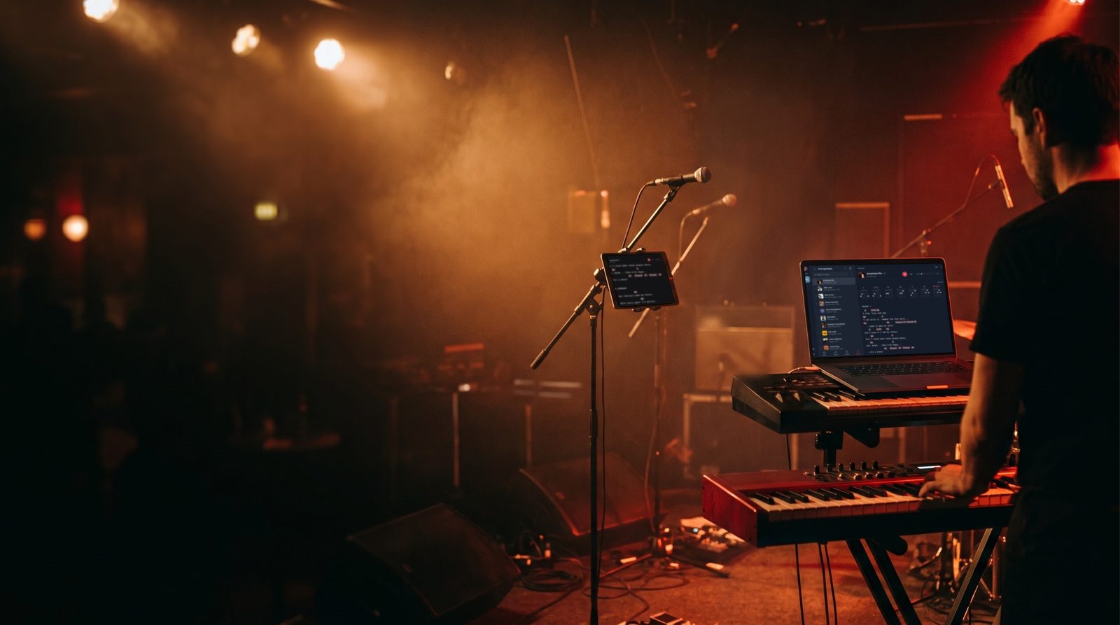 A keyboard player at soundcheck on a club stage reads Stage Friend on a laptop on his keyboard stand, beside a microphone stand with a tablet showing the same songs.