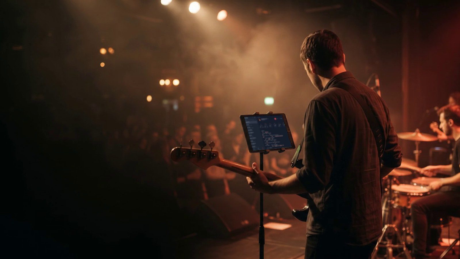 A bass player on a club stage, seen from behind, reads the band's setlist and the current song's chords in Stage Friend on a tablet on a stand, with the drummer and the crowd behind.