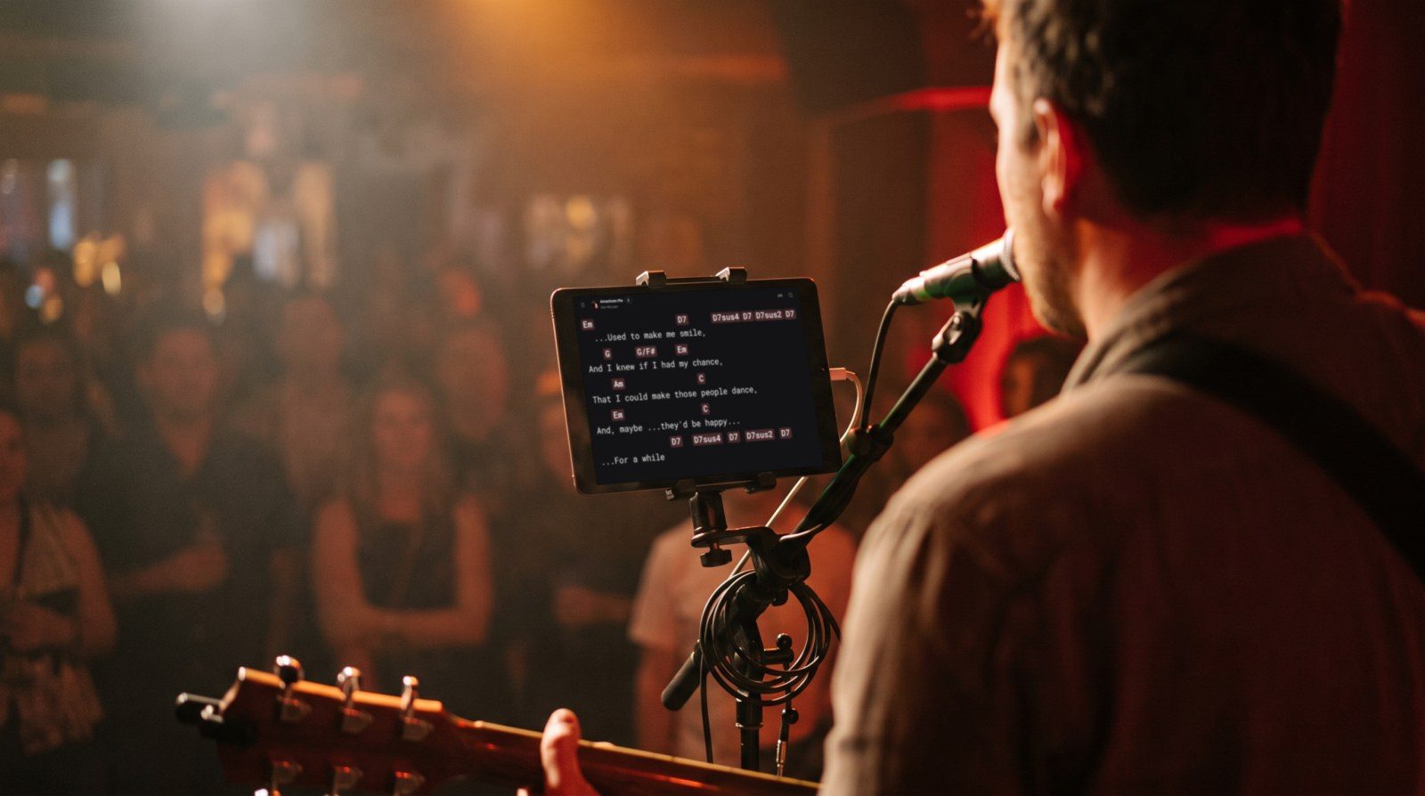 A singer with an acoustic guitar on a bar stage, seen from behind, reads lyrics and chords in Stage Friend on a tablet clamped to his microphone stand.