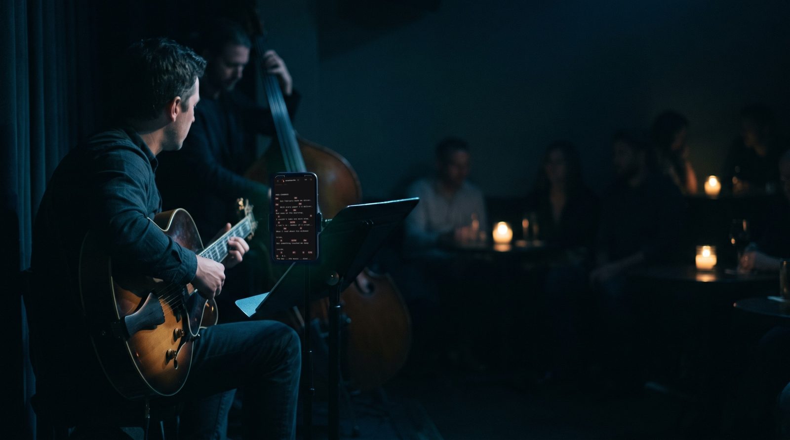 In a dim jazz bar late at night, a guitarist plays beside a music stand with a phone showing lyrics and chords in Stage Friend's near-black Stage theme.