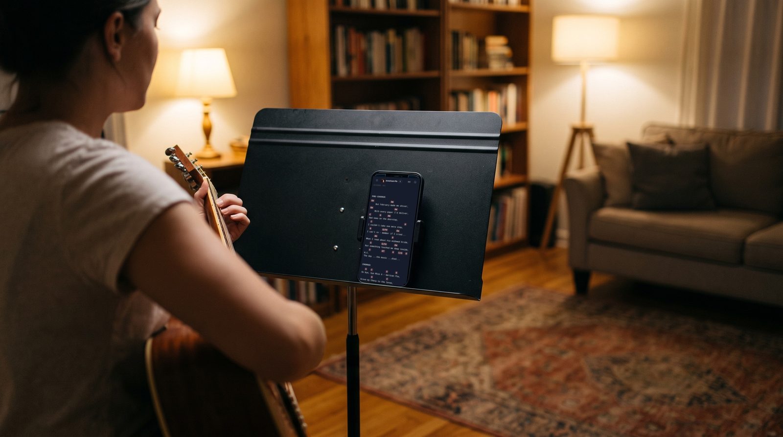 At home in the evening, someone practising alone sings and plays an acoustic guitar, seen from behind, reading the lyrics and chords of American Pie in Stage Friend on a phone clipped to a music stand.